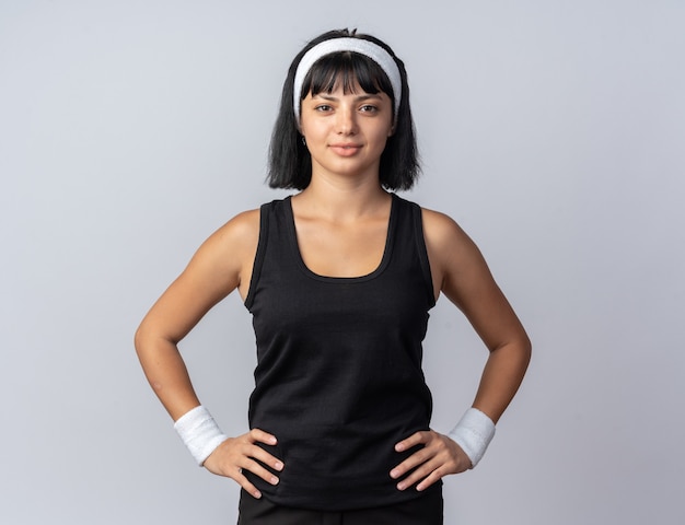 Giovane ragazza fitness che indossa la fascia guardando la telecamera con espressione seria e sicura con le braccia all'anca