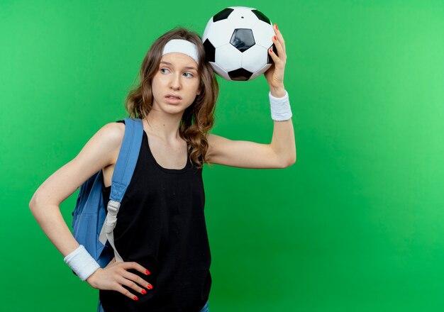 Giovane ragazza fitness in abiti sportivi neri con zaino e fascia tenendo il calcio sopra la testa lookign da parte perplesso in piedi sopra la parete verde