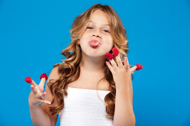 Giovane ragazza graziosa che mangia lampone sopra la parete blu