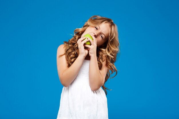 Giovane ragazza graziosa che mangia mela verde sopra la parete blu