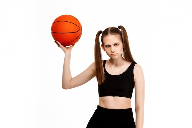 Giovane ragazza graziosa che propone con la pallacanestro, isolata sulla parete bianca