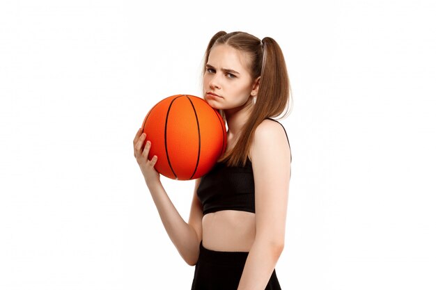 Giovane ragazza graziosa che propone con la pallacanestro, isolata sulla parete bianca