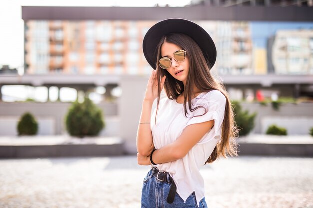 Giovane ragazza graziosa con black hat e occhiali da sole all'aperto sulla via soleggiata