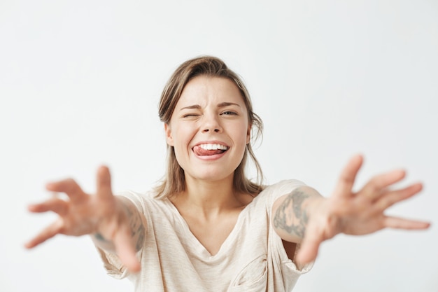 Giovane ragazza graziosa divertente che sbatte le palpebre mostrando la lingua che allunga le mani alla macchina fotografica.