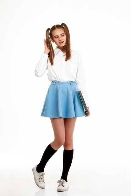 Giovane ragazza graziosa emotiva in uniforme in posa sul muro bianco