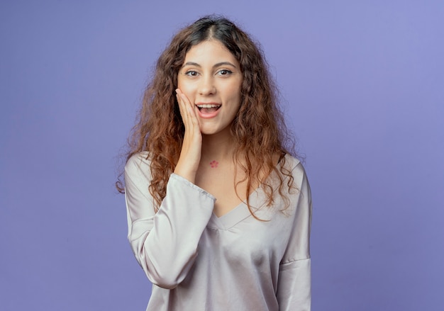 Giovane ragazza graziosa impressionata che mette la mano sulla guancia isolata su fondo blu