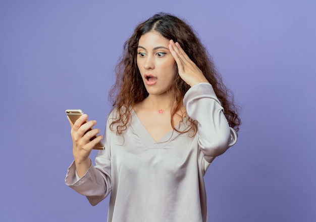 Giovane ragazza graziosa spaventata che tiene e che esamina il telefono e che mette la mano sulla fronte isolata sulla parete blu