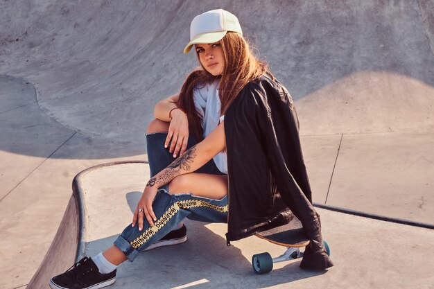 Giovane ragazza hipster in berretto vestita con jeans strappati e camicia bianca seduta su longboard allo skatepark.