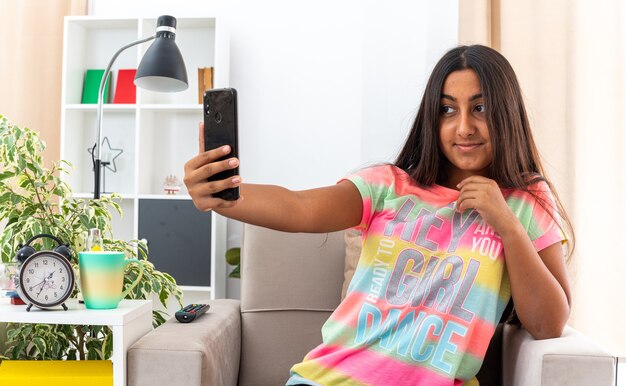 Giovane ragazza in abiti casual che fa selfie utilizzando lo smartphone sorridendo felice e positivo allegramente seduta sulla sedia in un soggiorno luminoso