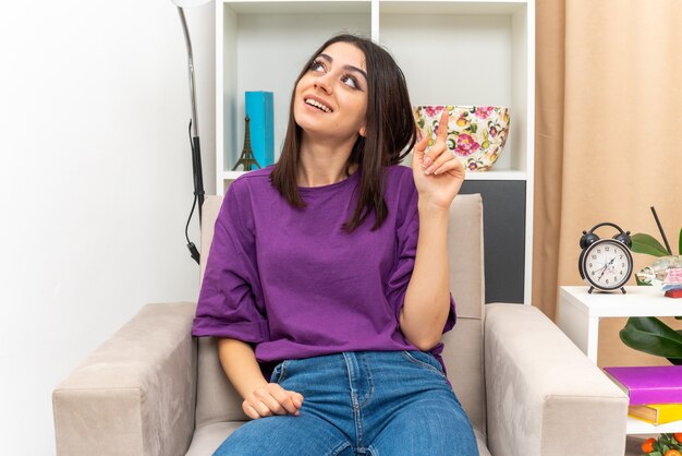 Giovane ragazza in abiti casual che guarda in alto con un sorriso sul viso felice e piositiva che mostra il dito indice seduto su una sedia in un soggiorno luminoso