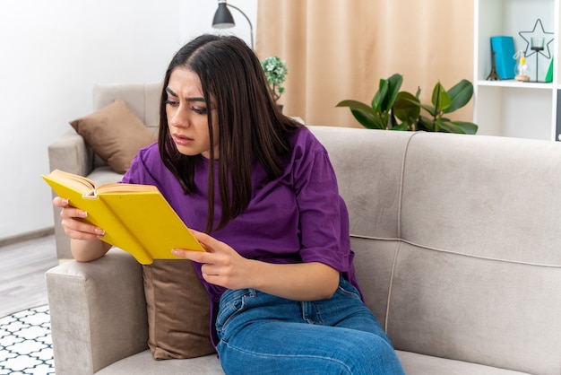 Giovane ragazza in abiti casual che legge un libro con una faccia seria seduta su un divano in un soggiorno luminoso