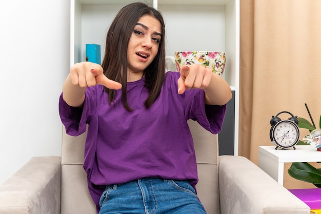 Giovane ragazza in abiti casual che sembra sicura di sé sorridente indicando con il dito indice seduto su una sedia in un soggiorno luminoso