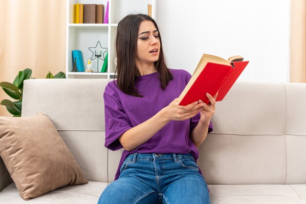 Giovane ragazza in abiti casual che tiene la lettura del libro con un'espressione confusa spesa weeked a casa seduto su un divano nella luce del soggiorno