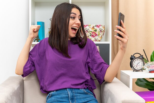 Giovane ragazza in abiti casual che tiene smartphone pugno serrato felice ed eccitato grida di gioia per il suo successo seduto su una sedia in soggiorno luminoso