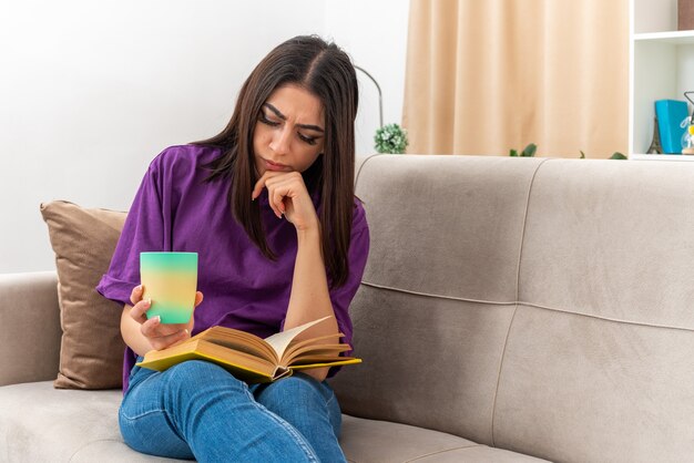 Giovane ragazza in abiti casual che tiene una tazza di tè leggendo un libro con una faccia seria seduta su un divano in un soggiorno luminoso