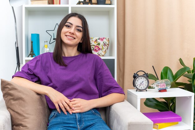 Giovane ragazza in abiti casual felice e positiva che sorride allegramente seduta su una sedia in un soggiorno luminoso