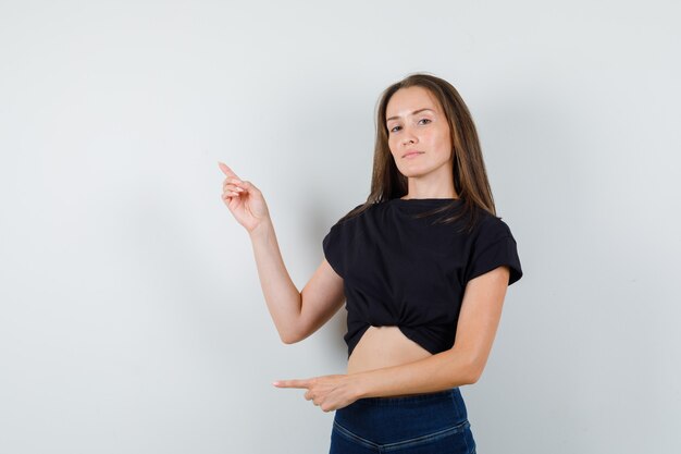 Giovane ragazza in camicetta nera, pantaloni che punta a lato e in alto e guardando positivo