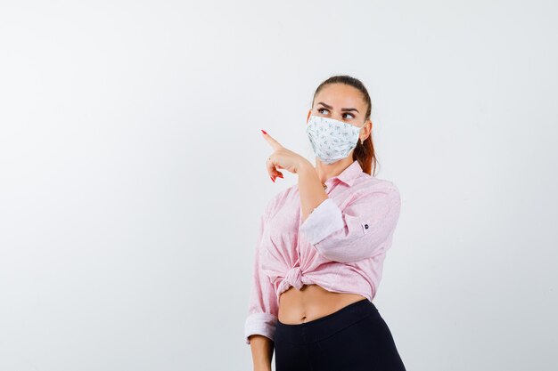 Giovane ragazza in camicetta rosa, pantaloni neri, maschera rivolta a sinistra con il dito indice, guardando lontano e seducente, vista frontale.