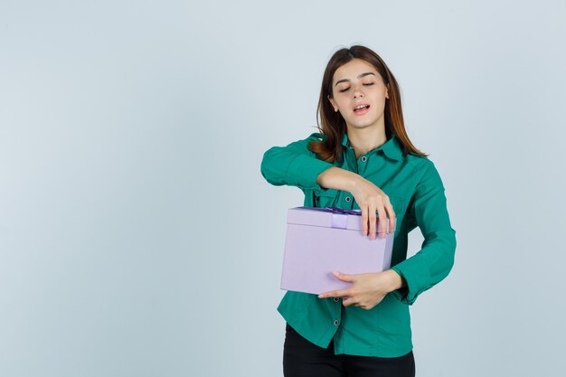 Giovane ragazza in camicetta verde, pantaloni neri, cercando di aprire la confezione regalo e guardando felice, vista frontale.