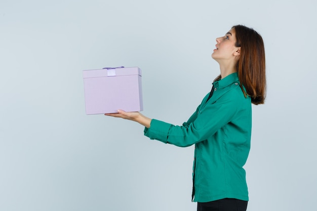 Giovane ragazza in camicetta verde, pantaloni neri che tiene confezione regalo, guardando verso l'alto e guardando concentrato, vista frontale.