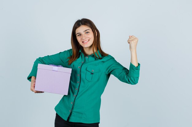 Giovane ragazza in camicetta verde, pantaloni neri che tiene confezione regalo, mostrando il gesto del vincitore e guardando fortunato, vista frontale