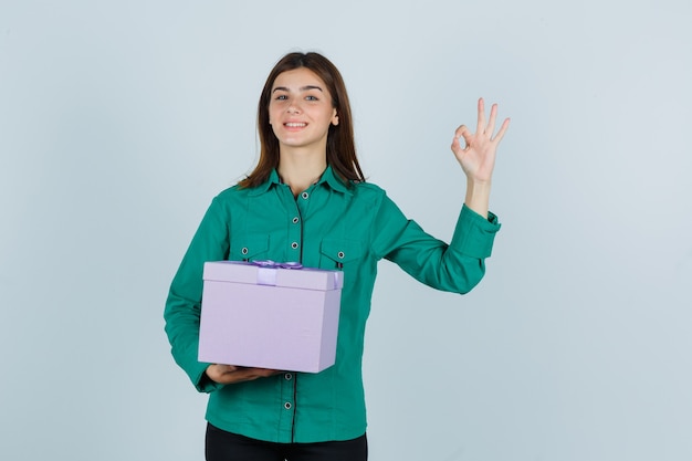 Giovane ragazza in camicetta verde, pantaloni neri che tiene confezione regalo, mostrando segno ok e guardando allegro, vista frontale.