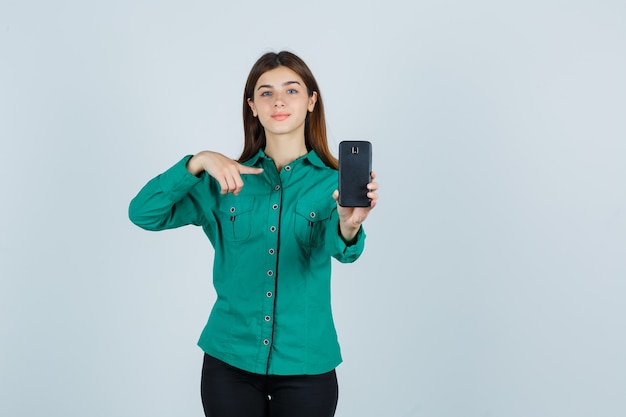 Giovane ragazza in camicetta verde, pantaloni neri che tiene il telefono in una mano, indicandolo con il dito indice e guardando allegro, vista frontale.