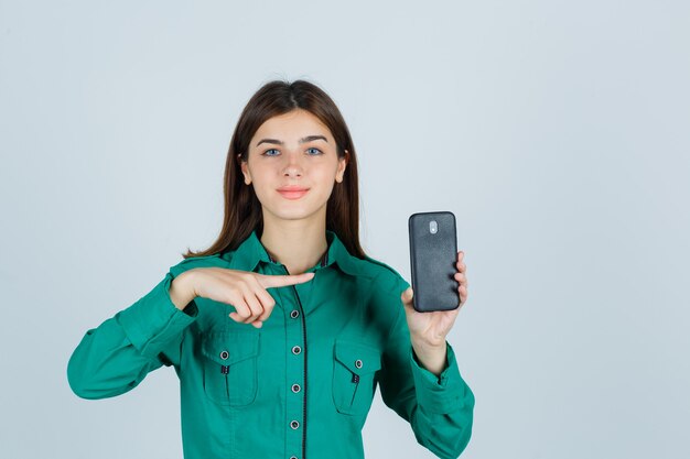 Giovane ragazza in camicetta verde, pantaloni neri che tiene il telefono in una mano, indicandolo e guardando allegro, vista frontale.