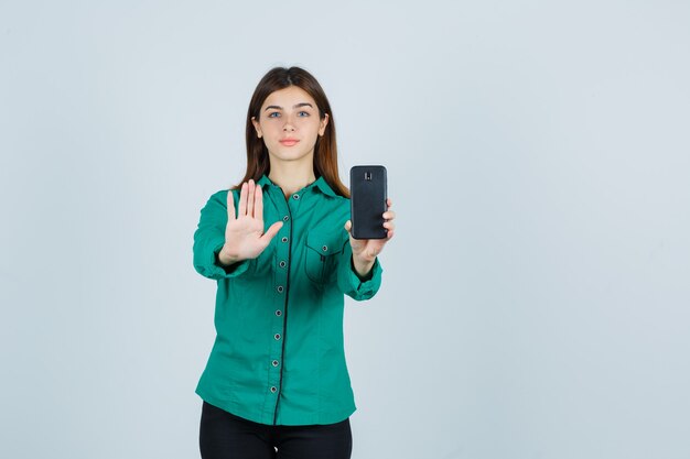 Giovane ragazza in camicetta verde, pantaloni neri che tiene il telefono in una mano, mostrando il segnale di stop e guardando serio, vista frontale.