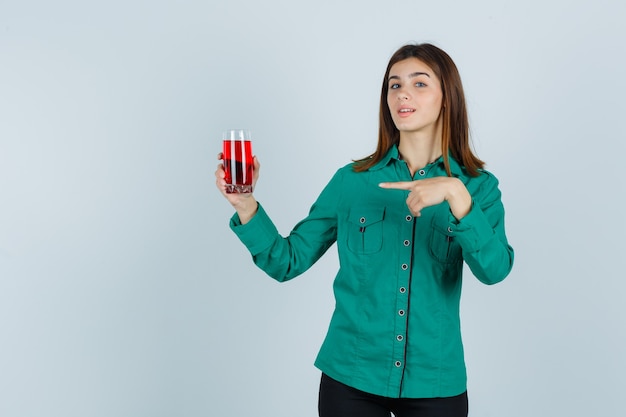 Giovane ragazza in camicetta verde, pantaloni neri con in mano un bicchiere di liquido rosso, indicandolo e guardando carino, vista frontale.