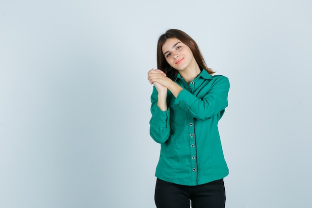 Giovane ragazza in camicetta verde, pantaloni neri stringendo le mani sul petto e guardando allegra, vista frontale.