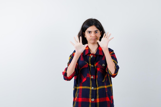 Giovane ragazza in camicia a quadri alzando i palmi in gesto di resa e guardando felice, vista frontale.