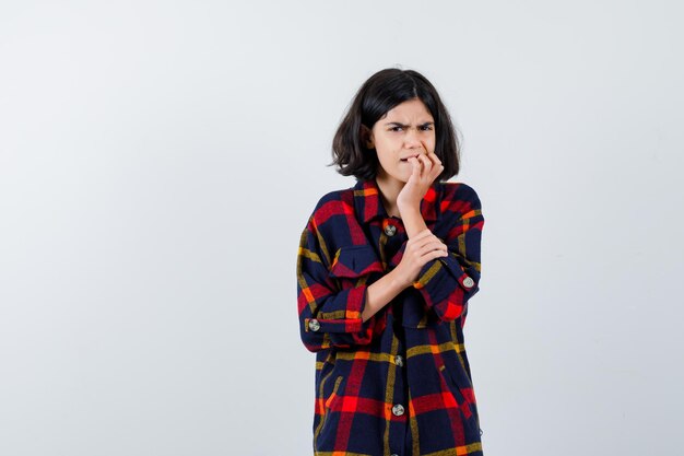 Giovane ragazza in camicia a quadri che morde le dita mentre tiene la mano sul braccio e sembra eccitata, vista frontale.