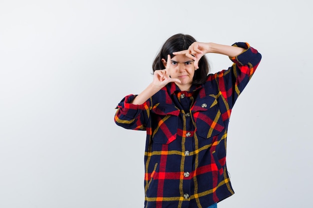 Giovane ragazza in camicia a quadri che mostra il gesto del telaio e sembra carina, vista frontale.