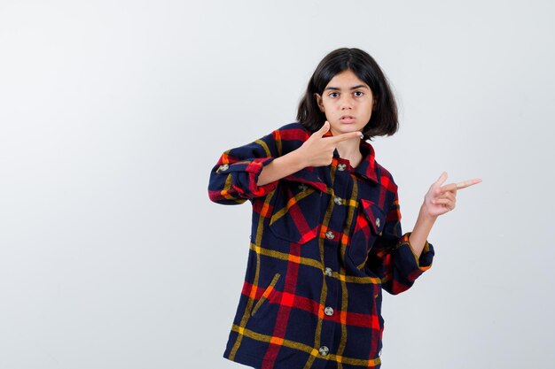 Giovane ragazza in camicia a quadri che punta a destra con l'indice e sembra seria, vista frontale.