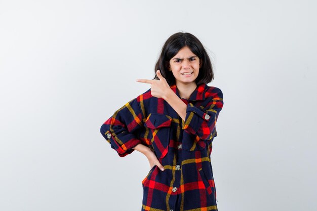 Giovane ragazza in camicia a quadri che punta a sinistra con il dito indice mentre tiene la mano sulla vita e sembra tormentata, vista frontale.