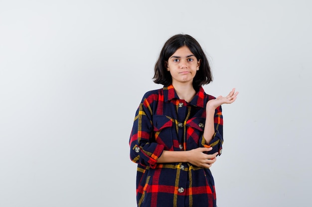 Giovane ragazza in camicia a quadri che tiene la mano sulla fronte mentre allunga la mano mentre tiene qualcosa e sembra seria, vista frontale.