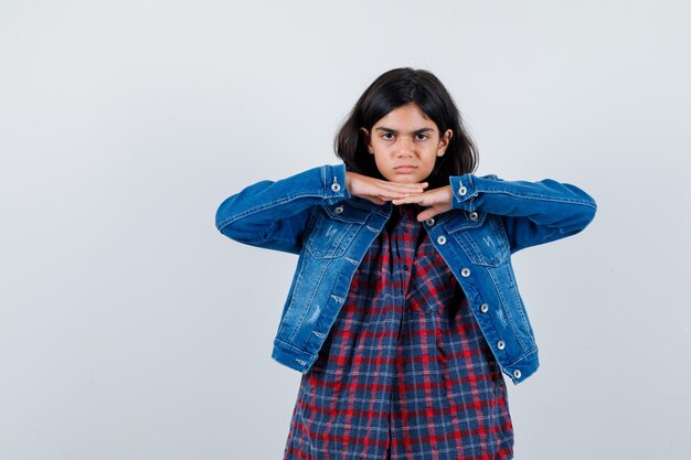 Giovane ragazza in camicia a quadri e giacca di jeans che si appoggia il mento sotto le mani e sembra seria, vista frontale.