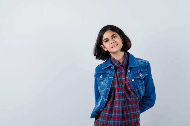 Giovane ragazza in camicia a quadri e giacca di jeans che si tiene per mano dietro la vita e sembra carina, vista frontale.