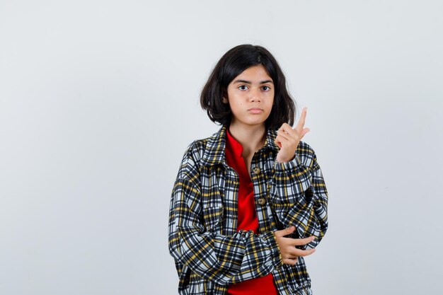 Giovane ragazza in camicia a quadri e t-shirt rossa che alza il dito indice nel gesto di eureka, tenendo la mano sul gomito e sembrando sensata, vista frontale.