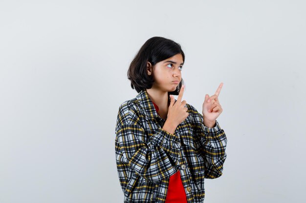 Giovane ragazza in camicia a quadri e t-shirt rossa che punta verso l'alto con l'indice e sembra seria, vista frontale.