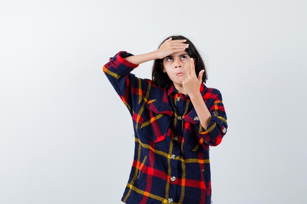 Giovane ragazza in camicia a quadri rivolta verso l'alto tenendo la mano sulla fronte e guardando pensierosa, vista frontale.