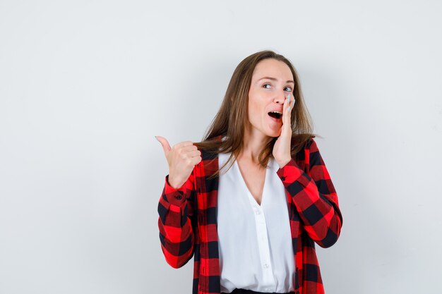 Giovane ragazza in camicia a scacchi, camicetta che dice il segreto, mostra pollice in su e sembra curiosa, vista frontale.