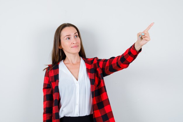 Giovane ragazza in camicia a scacchi, camicetta che punta verso l'alto con il dito e sembra soddisfatta, vista frontale.