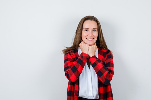 Giovane ragazza in camicia a scacchi, camicetta che tiene i pugni sotto il mento e sembra soddisfatta, vista frontale.