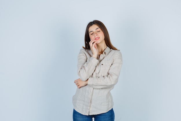 Giovane ragazza in camicia beige, jeans appoggiati alla guancia, tenendo gli occhi chiusi e guardando affascinante, vista frontale.