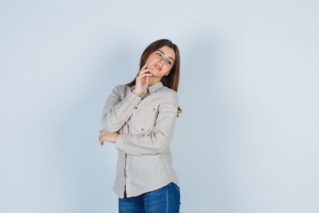 Giovane ragazza in camicia beige, jeans che mette la mano sotto il mento, distoglie lo sguardo e sembra carina, vista frontale.