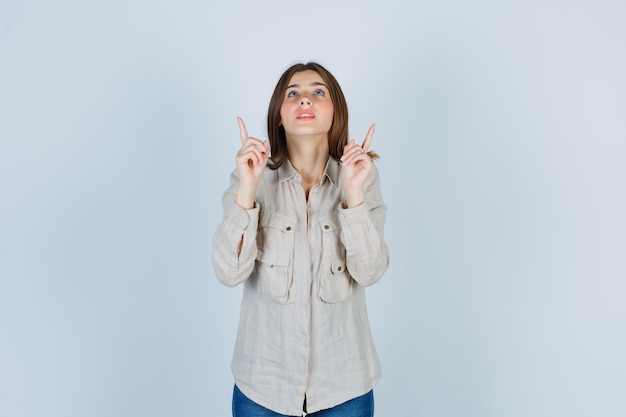 Giovane ragazza in camicia beige, jeans rivolti verso l'alto con l'indice e guardando concentrati, vista frontale.