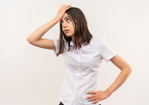 Giovane ragazza in camicia bianca che osserva da parte confusa con la mano sulla sua testa per errore in piedi sul muro bianco