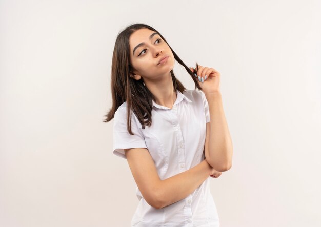 Giovane ragazza in camicia bianca che osserva da parte in piedi perplesso sopra il muro bianco